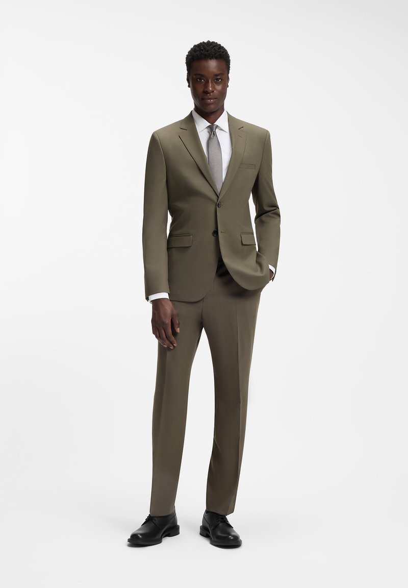 Mann posiert in olivgrünem Anzug mit weißem Hemd, grauer Krawatte und schwarzen Lederschuhen vor weißem Hintergrund.