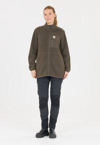 Giacca in fleece marrone con chiusura a zip, due tasche frontali e dettaglio a patch. Abbinata a pantaloni scuri con tasche con zip e sezioni testurizzate.
