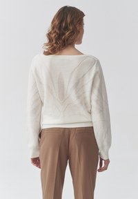 Suéter de punto blanco con patrón floral, amplio escote barco, puños y dobladillo acanalados. Combinado con pantalones de sastre marrones.