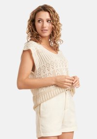 Beige gebreid top met een V-hals, voorzien van een open, geometrisch patroon. Gecombineerd met bijpassende beige shorts, die een gestructureerde stof laten zien.