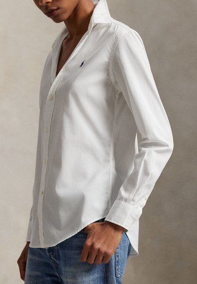 Persona que lleva una camisa blanca de manga larga con botones y un pequeño logo bordado, y vaqueros azules, de pie frente a un fondo neutro.