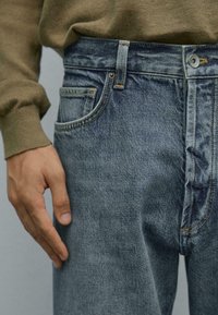 Primo piano di una mano di una persona che riposa vicino alla tasca anteriore di un paio di jeans blu sbiaditi e indossa un maglione marrone a maniche lunghe.