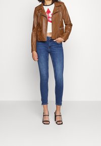 Blouson de moto en cuir marron avec ferrures argentées, haut court blanc avec un motif rouge, jean skinny bleu et talons noirs à lanières.