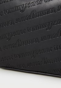 Svart läderpås med präglat varumärke "tommyjeans" i ett upprepat mönster. Slät yta, rektangulär form, minimalistisk design.