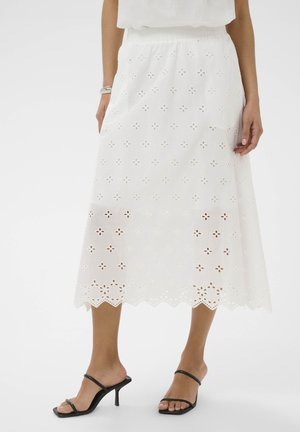 Kobieta ubrana w białą spódnicę midi z ażurowymi kwiatowymi wycięciami, z falbaniastym dołem, srebrną bransoletkę i czarne sandały na obcasie z paskami.
