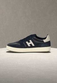 Μπλε ναυτικό και μαύρο αθλητικό παπούτσι με άσπρη σόλα και ταμπελάκι στη φτέρνα, με μεγάλο άσπρο γράμμα "H" στο πλάι, τοποθετημένο σε ανοιχτό γκρι επιφάνεια.