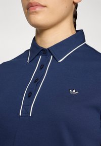 Marineblauwe poloshirt met een effen textuur, witte kraagafwerking en drie knopen. Heeft een klein geborduurd logo op de linkerborst.