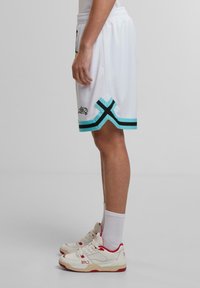 Weiße Basketballshorts mit schwarzen und türkisfarbenen Akzenten, ausgestattet mit einem elastischen Bund und einem geometrischen Saumdesign. Kombiniert mit weißen und roten Turnschuhen.