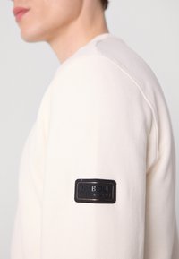Creme-kleurige sweatshirt met lange mouwen en een gestructureerde stof, voorzien van een zwart leren label op de linkermouw met het merk "BARBOUR INTERNATIONAL."