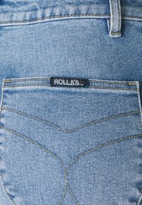Jeans de ganga azul clara com um design de bolso em forma de chevron distinto, apresentando costuras amarelas e um rótulo preto que diz "ROLLAS."