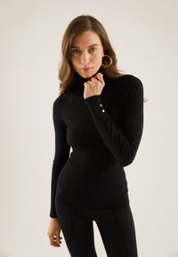 Svart ribbestrikket høyhalset topp med lange ermer, med gullknapper ved mansjettene, tettsittende design og myk tekstur.