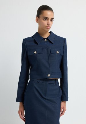 Donna che indossa un blazer blu navy su misura con bottoni dorati e una gonna alta coordinata, in piedi davanti a uno sfondo bianco uniforme.