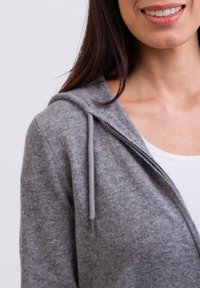 Grauer Reißverschluss-Hoodie aus weichem Material mit Kapuze. Mit einem silbernen Reißverschluss und Kordelzügen, über einem weißen Oberteil getragen.