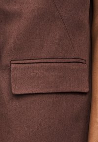 Καφέ υφασμάτινη τσέπη με καπάκι σε ένδυμα, που δείχνει λεπτομερή ραφή και μέρος του μανικιού στη δεξιά πλευρά.