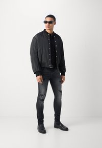 Cazadora bomber negra con una textura suave, con botones a presión, combinada con jeans oscuros desgastados y zapatos negros. Unas gafas de sol rectangulares completan el look.
