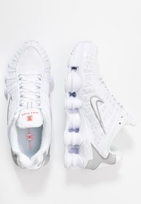 Vita nätfötter med grå detaljer och en framträdande silverfärgad Nike-swoosh. Har en distinkt vadderad sula och texturerade mönster.