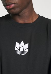 Svart bomull t-shirt med rund halsringning som har en vit broderad logotyp med tre löv och tre ränder på bröstet.