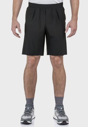Mann trägt schwarze knielange Shorts, graue Sportsocken und graue Laufschuhe, steht mit entspannten Armen an den Seiten.