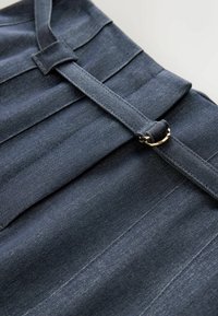 Produit en denim foncé avec un design plissé, doté d'une ceinture en tissu avec un accent matériel à anneau doré. Texture lisse et forme structurée.