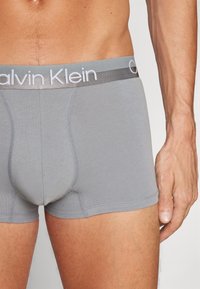 Pilki medvilniniai apatiniai šortai su minkštu juosmens dirželiu, ant kurio yra "Calvin Klein" logotipas, sklandžiu paviršiumi ir prigludusia konstrukcija su formuojančia kišenėle.