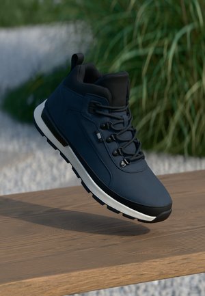 Navy blue mid-top shoe with black accents, featuring a lace-up design, textured material, and a white rubber sole. Mounted on a wooden surface.