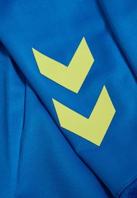 Blauwe stof met twee gele chevronpatronen. Het materiaal heeft een textuur, wat een sportief ontwerp benadrukt met gladde en geribbelde secties.