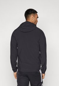 Giacca nera con cappuccio, maniche lunghe e polsini elastici, caratterizzata da un design semplice e da una texture leggera e liscia.