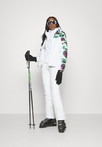Skioutfit som inkluderar en vit jacka med blommig ärmar, vita byxor, svarta handskar och skidstövlar, med skistavar i handen.