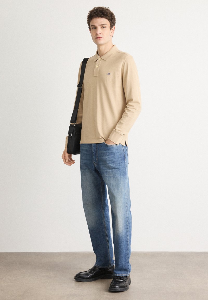 Beige Langarm-Polohemd mit Kragen, blaue Jeans und schwarze Schuhe. Das Model hält eine kleine schwarze Umhängetasche. Einfaches Design und lässige Passform.