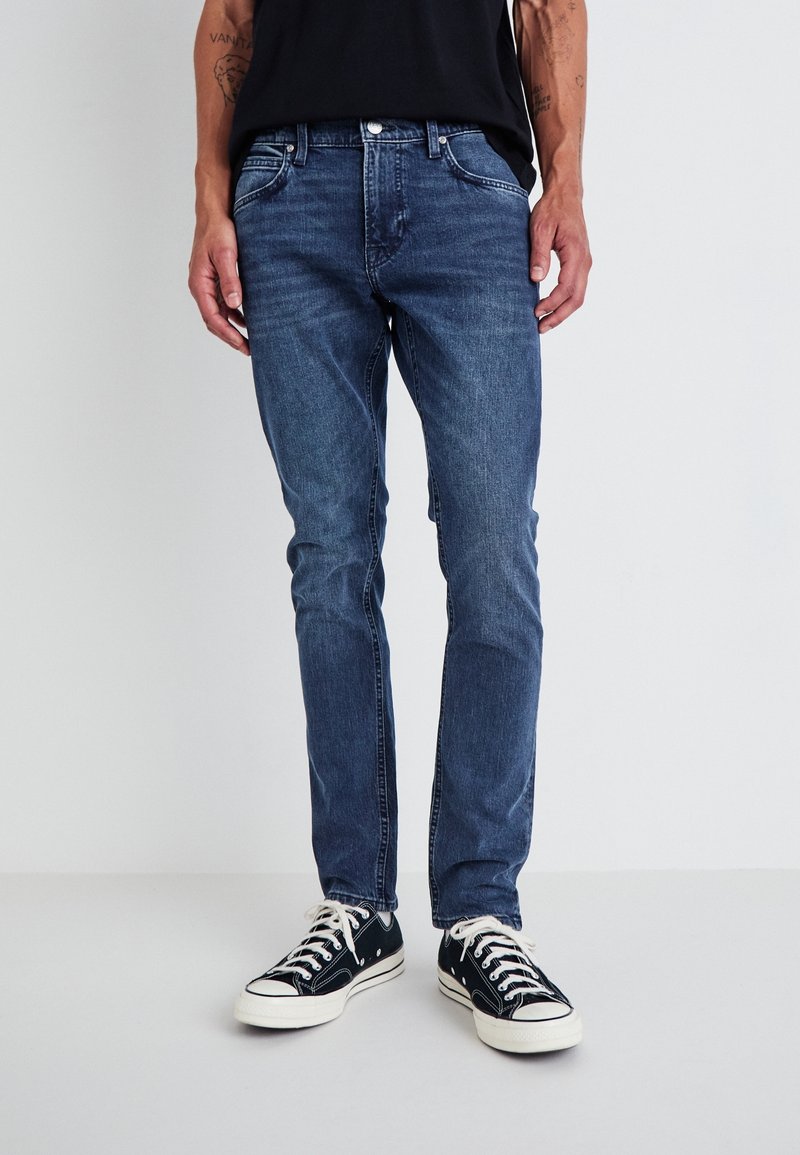 Jean en denim bleu à coupe slim, cinq poches, délavage subtil et coutures contrastées, assorti à des baskets en toile noire.