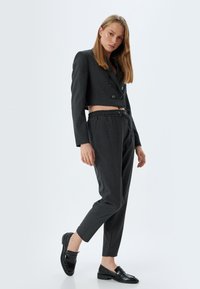 Grijze cropped blazer met dubbele knopen, gecombineerd met grijze drawstring broek en glanzende zwarte loafers, geplaatst op een neutrale achtergrond.