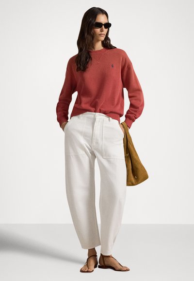 Rdeč pulover, bele široke hlače in rjava zamsasta torba. Model nosi črna sončna očala in bež sandale, izražajoč sproščen, sodoben videz.