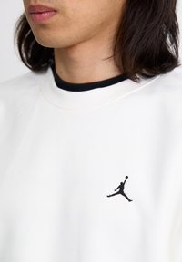 Hvid sweatshirt med en sort ribbet halsudskæring, prydet med et lille sort Jumpman-logo på brystet. Blød stoftekstur.