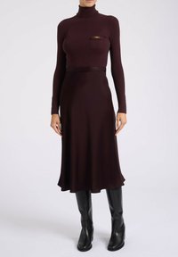 Haut en tulle côtelé bordeaux avec une poche poitrine et un accent doré, associé à une jupe fluide bordeaux et des bottes noires jusqu'aux genoux.