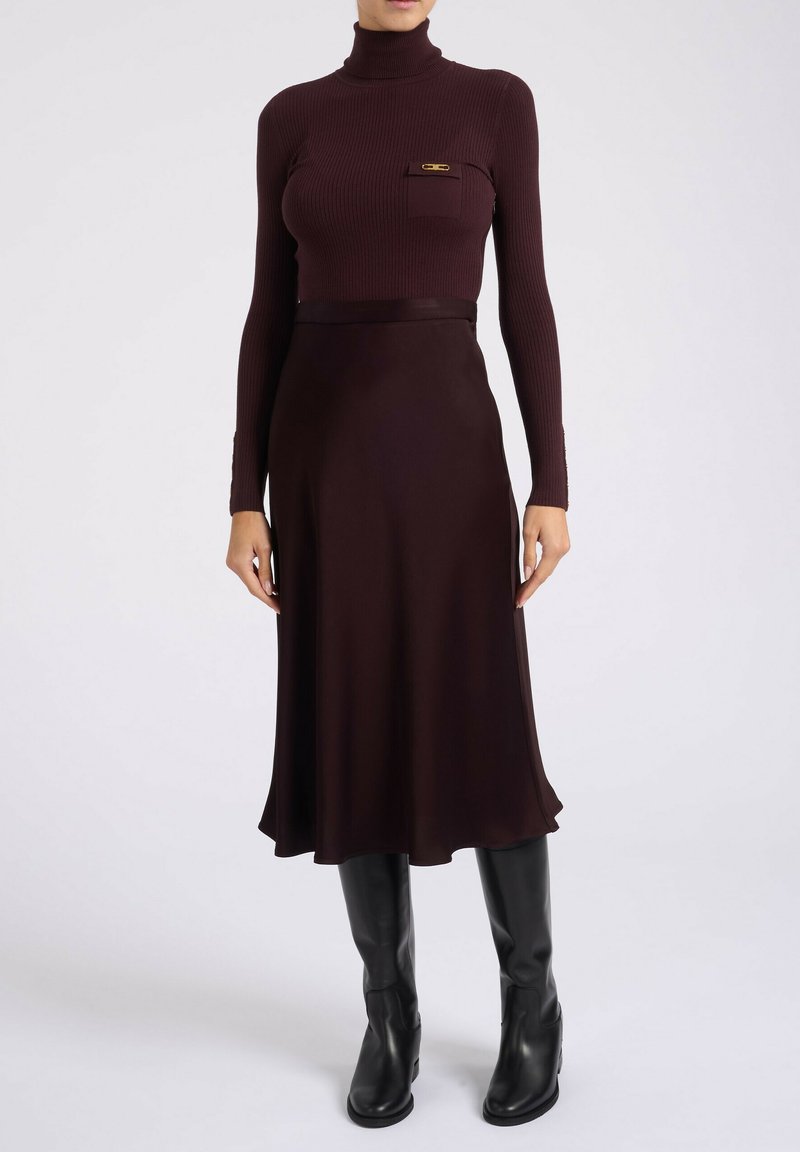 Haut en tulle côtelé bordeaux avec une poche poitrine et un accent doré, associé à une jupe fluide bordeaux et des bottes noires jusqu'aux genoux.