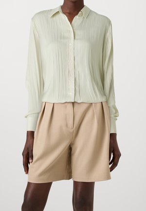 Osoba ubrana w jasnooliwkową koszulę na guziki z długim rękawem i pionowymi plisami oraz beżowe, wysokie, plisowane szorty.