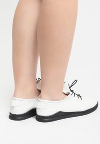 Zapatos blancos perforados con cordones y suela negra. Con puntera redondeada, cordones negros y una pestaña en el talón para ponerlos y quitarlos fácilmente.