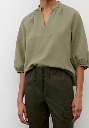 Femme portant un chemisier ample vert olive avec des manches ballon trois-quarts et un pantalon taille haute vert foncé avec un détail de poche latérale.