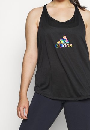 Vrouw die een mouwloos zwart Adidas-topje met een kleurrijk geometrisch logo en marineblauwe leggings draagt, staand tegen een effen achtergrond.