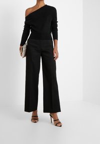 Žena ve černém svetr s jedním ramínkem a širokými kalhotami, držící béžovou kabelku, stojící ve bílém studiu, obutá v černých sandálech na vysokém podpatku.