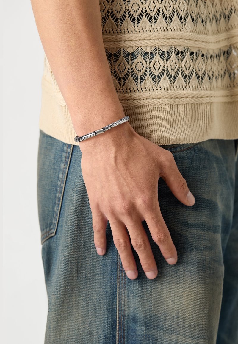 Main droite portant un bracelet en argent texturé, reposant à côté d'un jean bleu délavé et d'un haut beige tricoté avec un motif en dentelle.