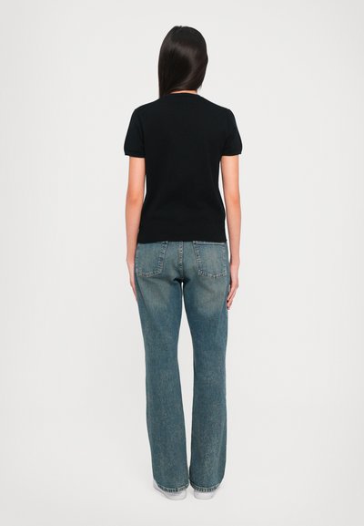 Woman with long dark hair wearing a black short-sleeve shirt, blue jeans, and white shoes, standing facing away on a plain background.
