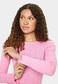 Vrouw met krullend haar die de mouw van een roze, gestreepte longsleeve blouse aanpast, terwijl ze naar links kijkt tegen een witte achtergrond.