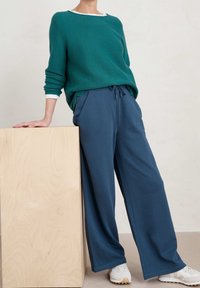 Grön bomullströja med ribbad struktur och vit halsringning, parat med löst sittande blå byxor och vita sneakers.