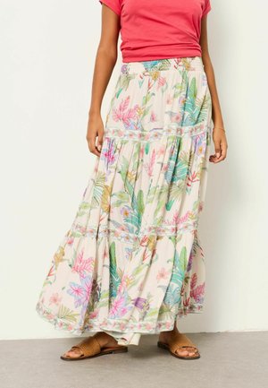 Frau trägt einen langen, fließenden Rock mit buntem tropischem Blumendruck, ein korallenfarbenes kurzärmeliges Oberteil und braune gewebte Sandalen und steht drinnen.