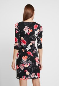 Zwarte bloemenjurk met lange mouwen, versierd met rode, roze en witte bloemen. Het materiaal lijkt zacht en rekbaar, met een aansluitend ontwerp.