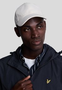 Vit bomullsbasketcap med en sydd krona och en böjd skärm. Kapseln bärs med en marinblå jacka och en randig skjorta under.