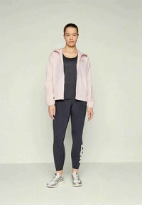 Mlada žena stoji, nosi svijetloružičastu jaknu na rajsferšlus, crnu majicu, crne tajice s bijelim logotipom i bijelo-sive tenisice, na jednostavnoj pozadini.