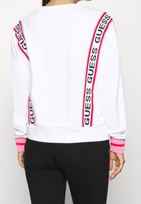 Sudadera blanca con rayas rosas y negras y la marca "GUESS". Tiene un cuello redondo y puños acanalados. La tela parece suave.