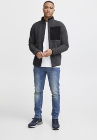 Chaqueta de forro polar gris con bolsillo negro, cremallera completa y cuello alto. Combinada con jeans azules y zapatillas azul marino. Diseño limpio y moderno.